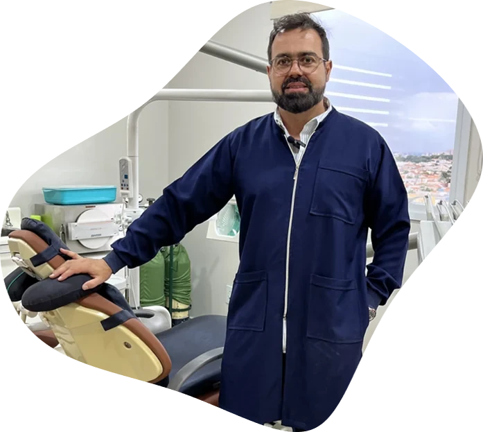 Dr. Paulo Melo em pé ao lado da cadeira, em seu consultório